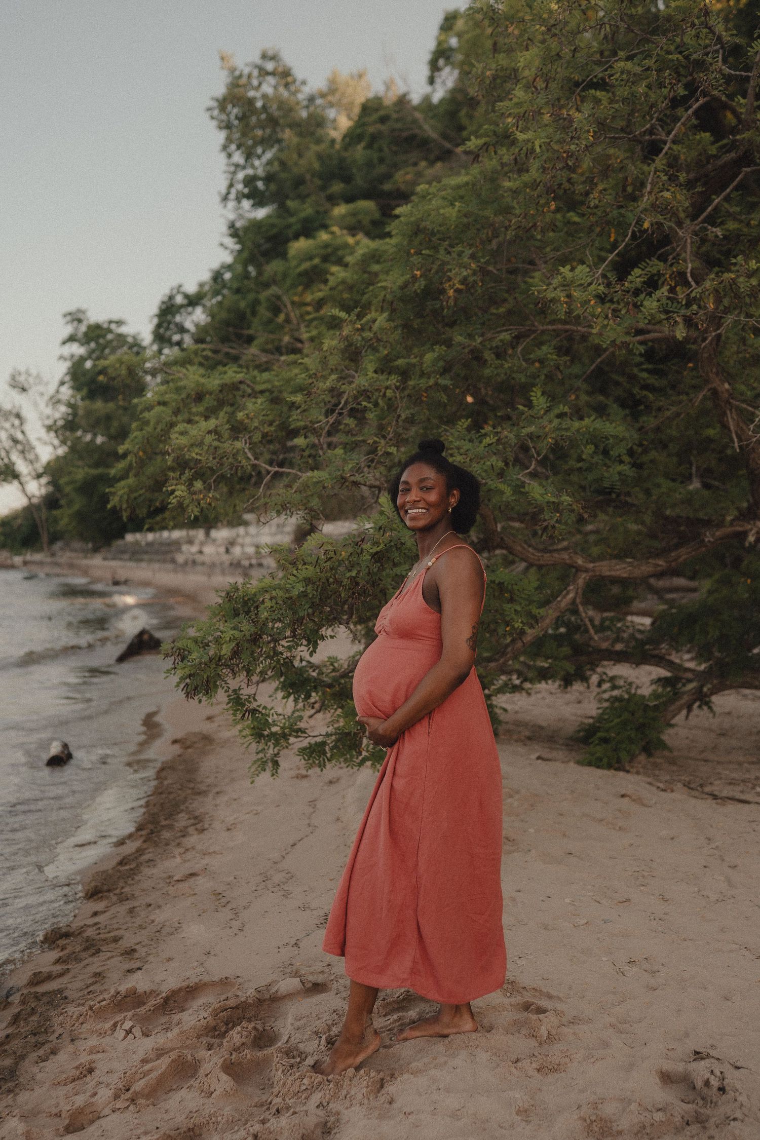 A smiling pregnant Black woman in a coral maxi dress stands barefoot on a sandy beach at dusk.