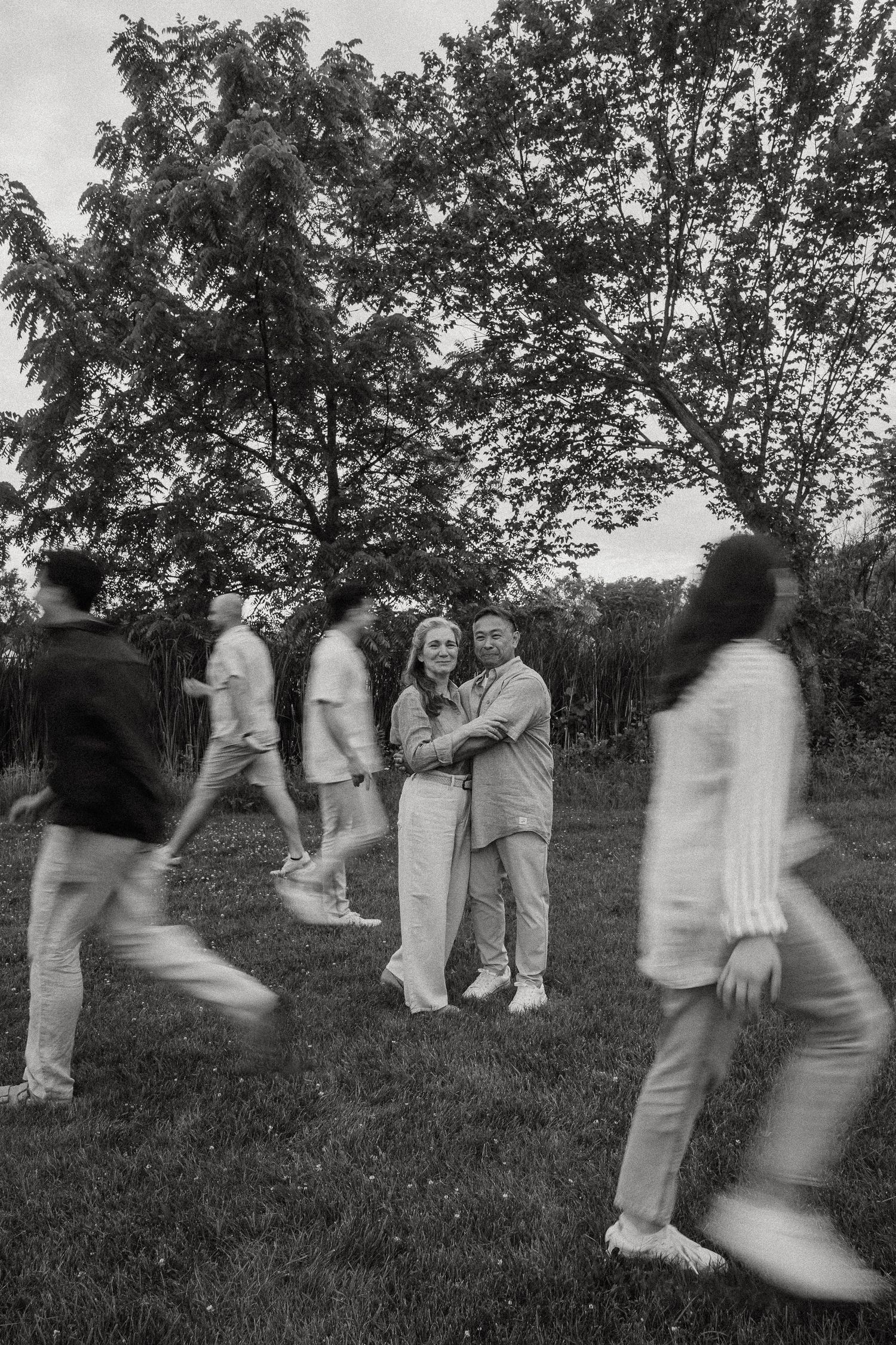 Black and white photo of a couple embracing in a park while blurred figures walk around them.