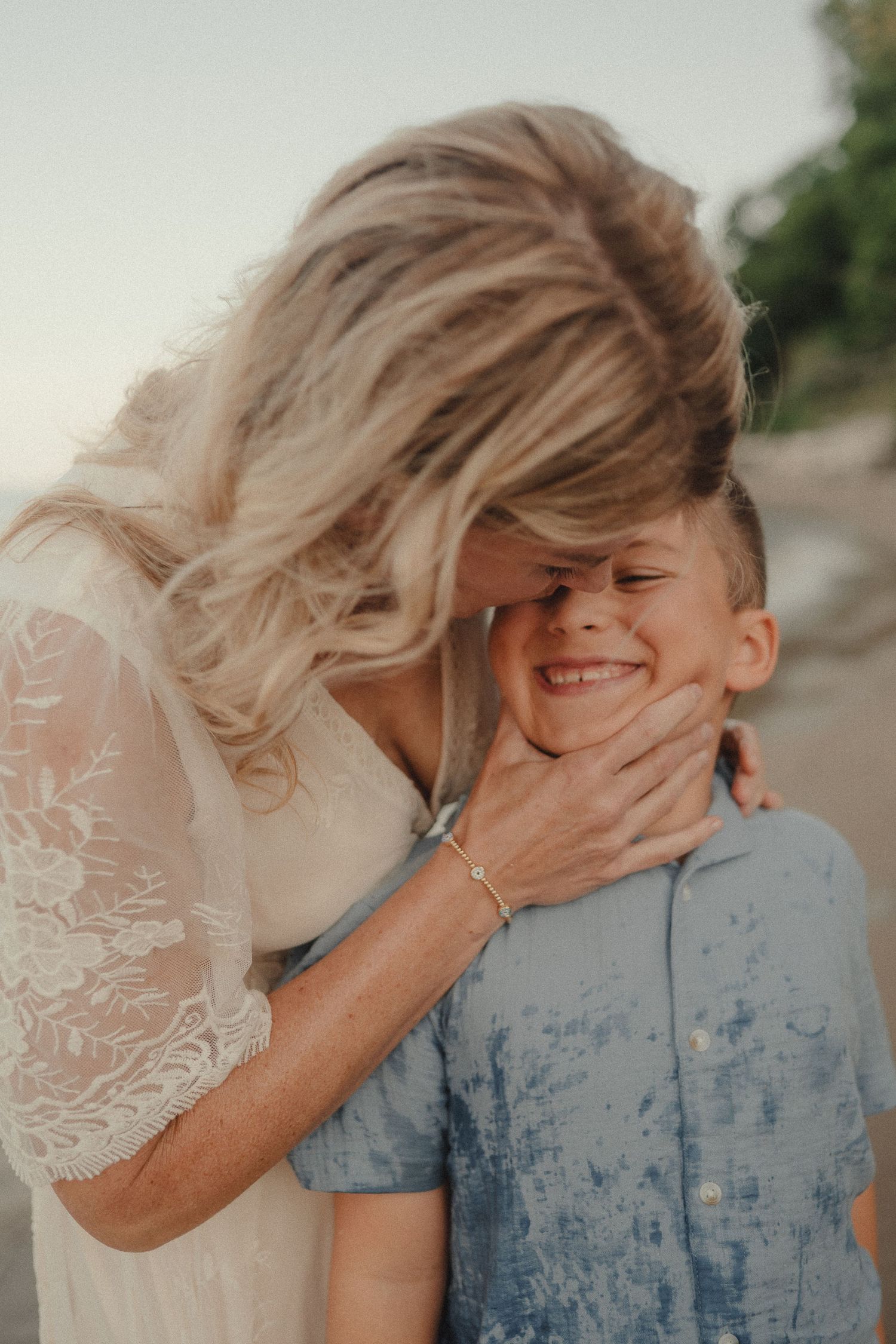 Laughing boy being affectionately embraced by his blonde mother on a breezy beach.