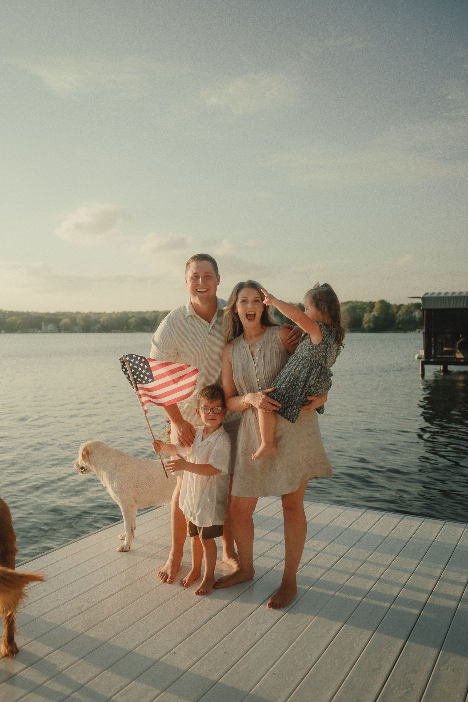 Happy family with dog and American flag posing on lake dock at sunset.