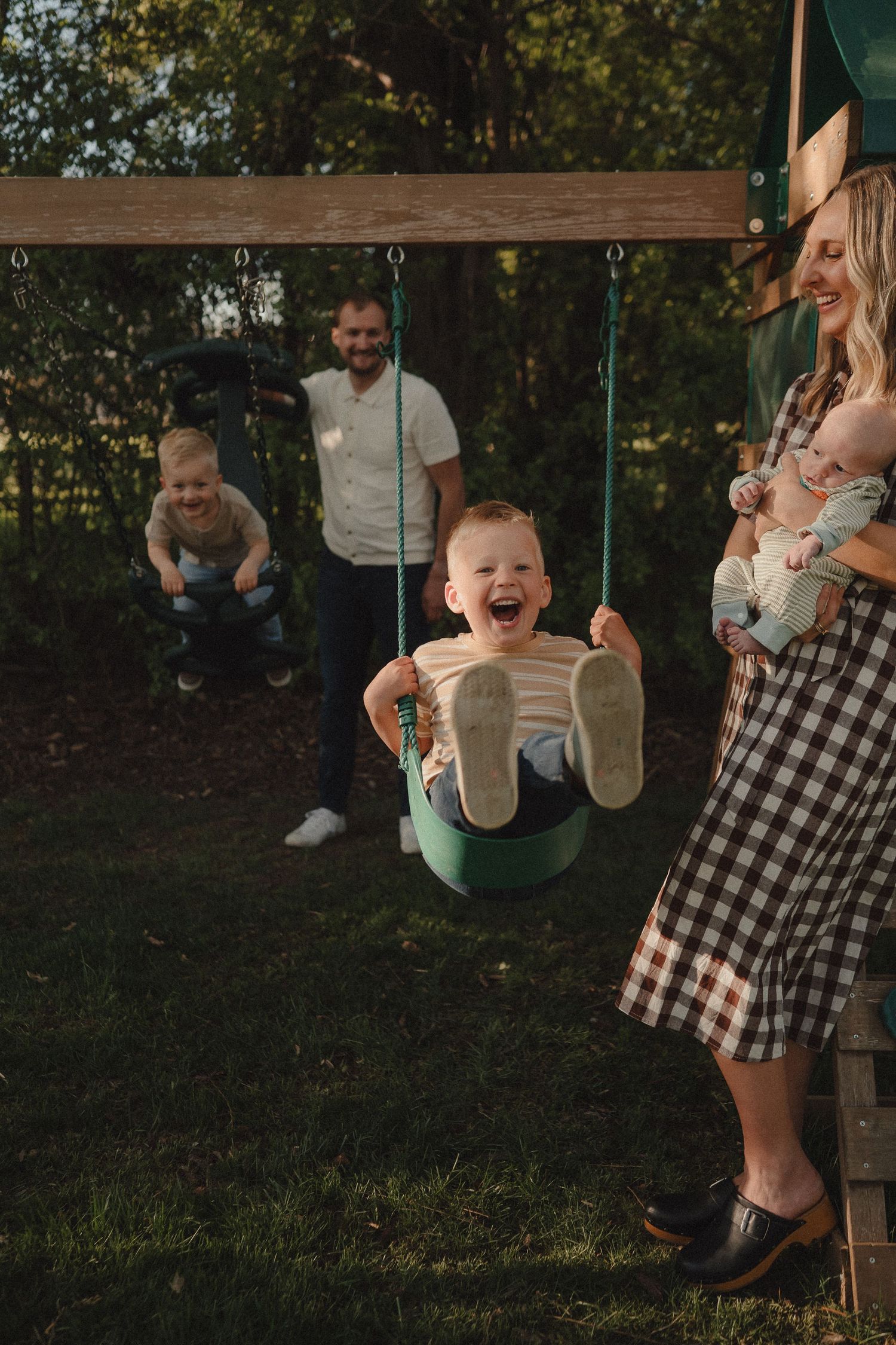 A joyful toddler laughs on a swing while his family plays outdoors on a sunny day.