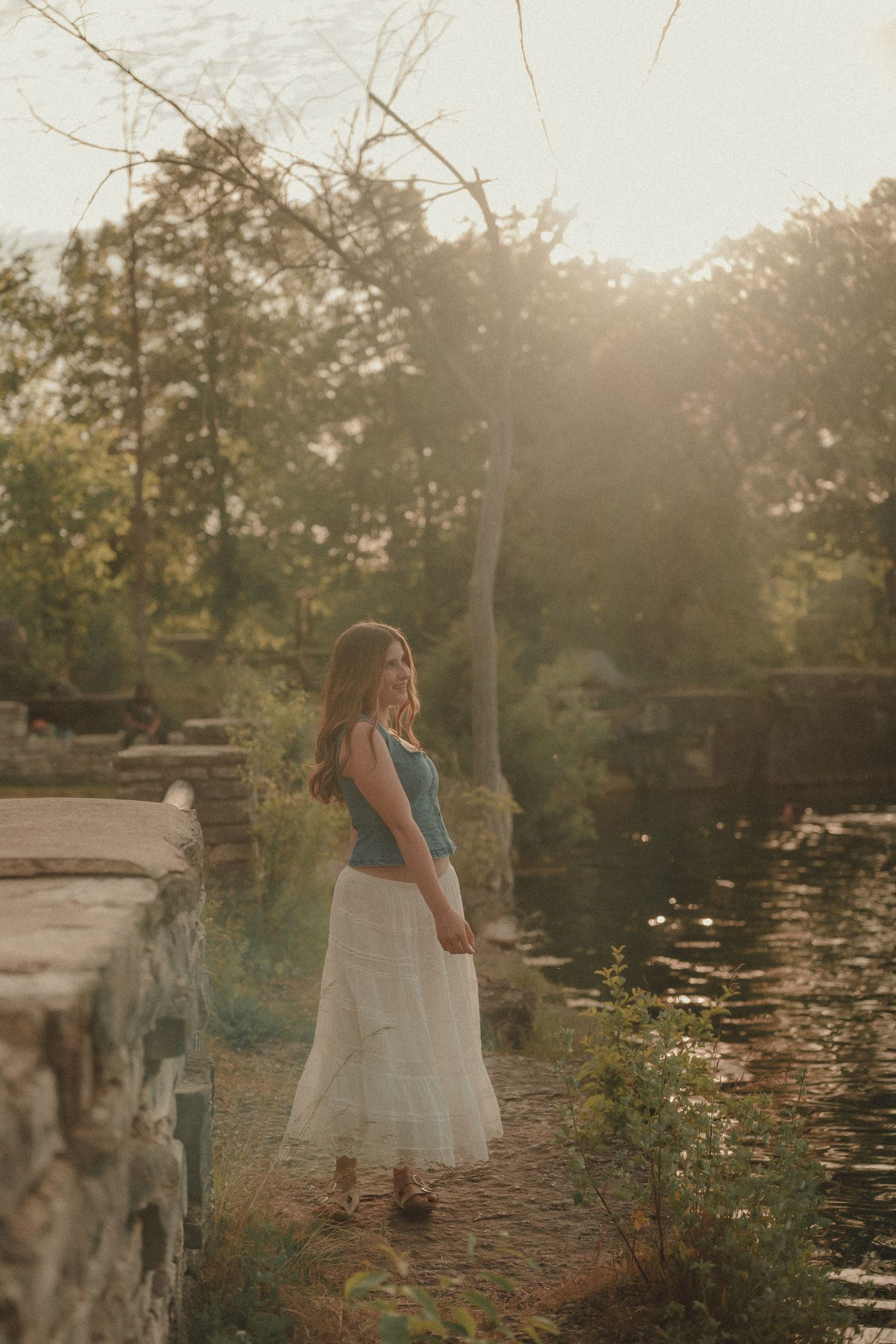 Young woman in white skirt and blue top stands by a sunlit pond surrounded by lush trees and stone walls.