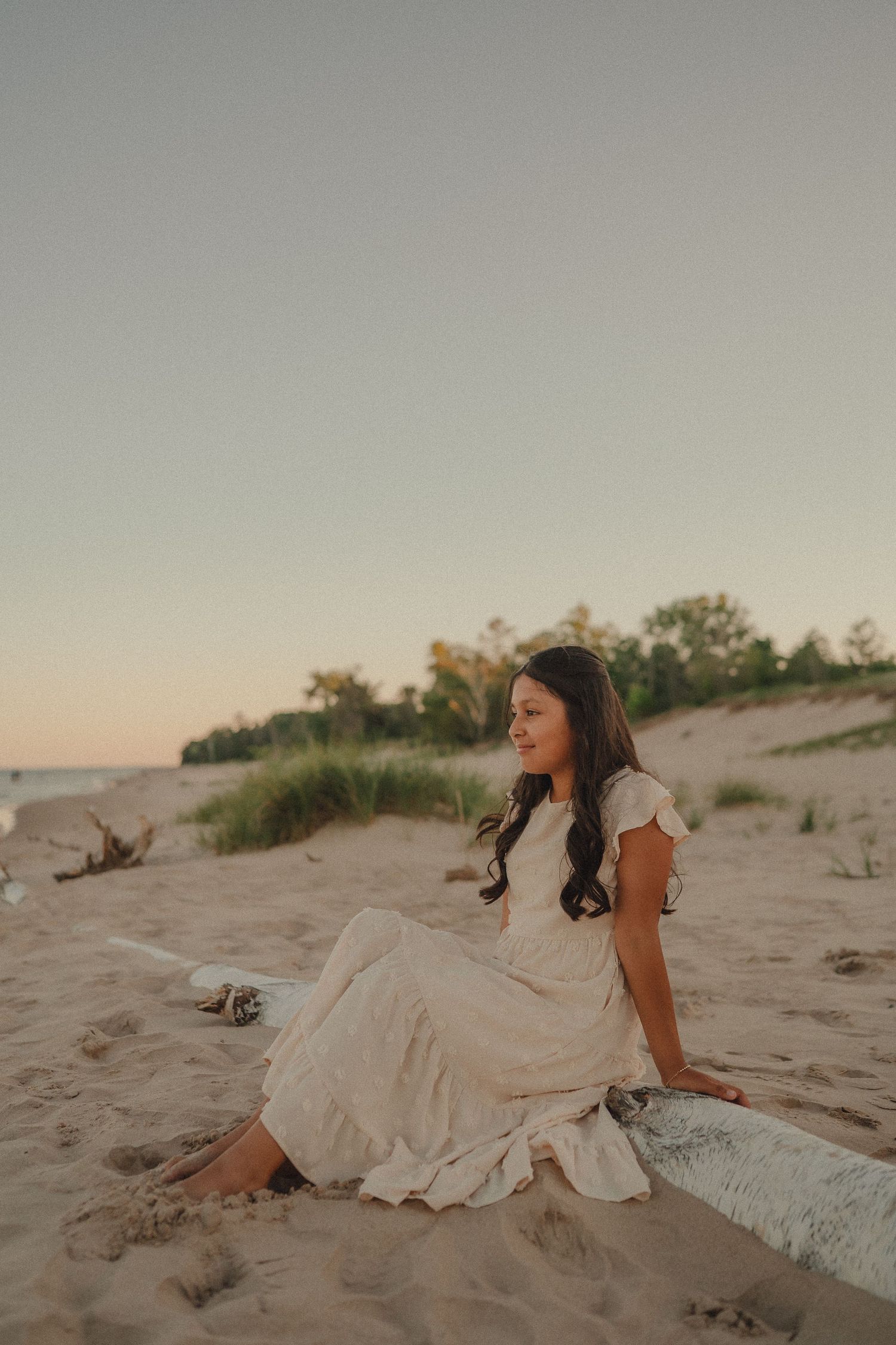 Young girl in white dress sits on driftwood on sandy beach at golden sunset.