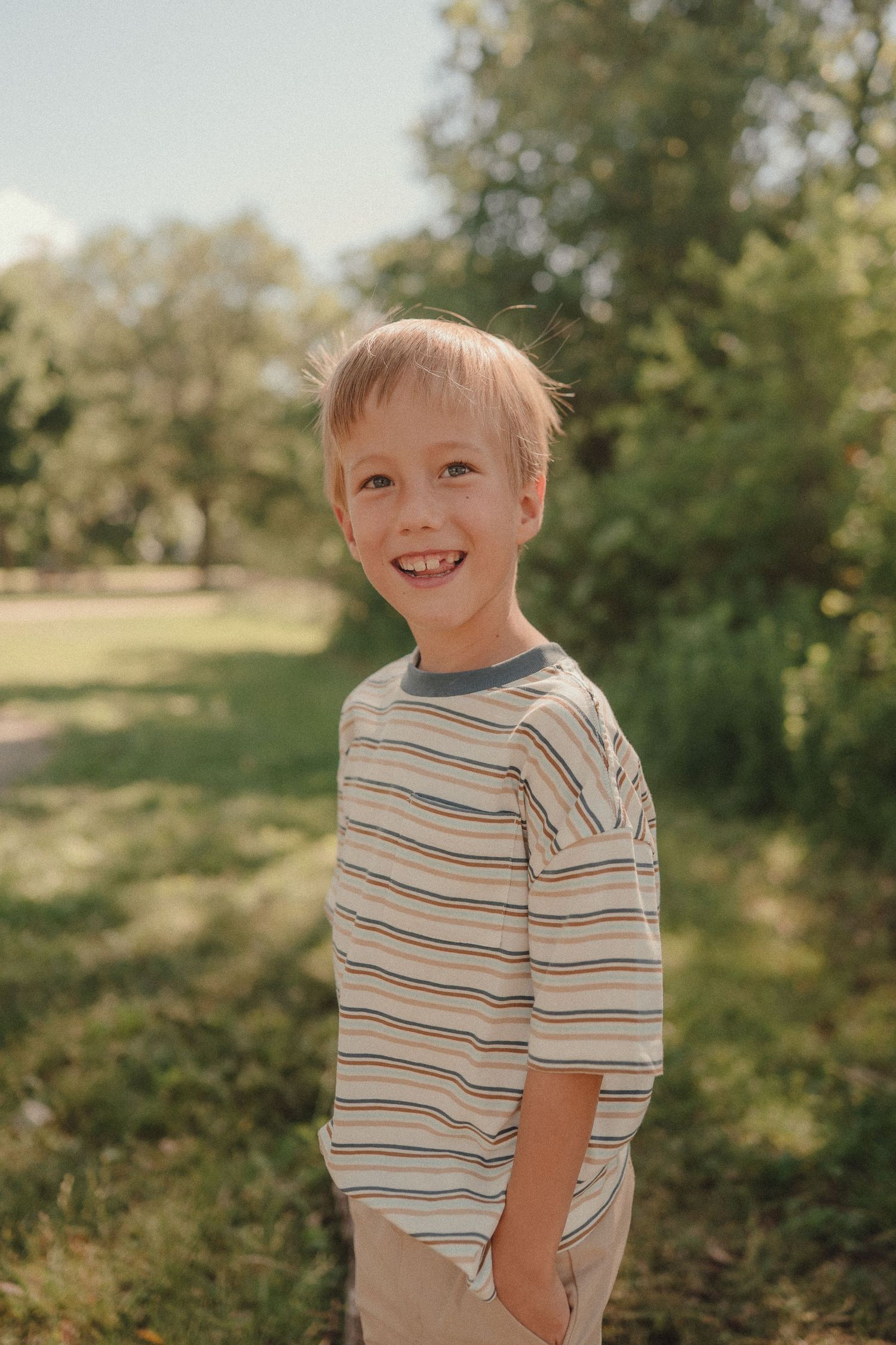 Smiling blonde boy in striped shirt standing outdoors in sunny green park setting.
