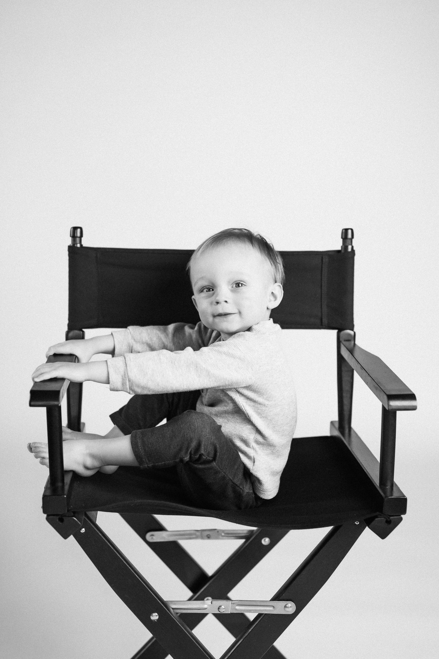Toddler in white shirt plays on black director's chair in black and white photo series.