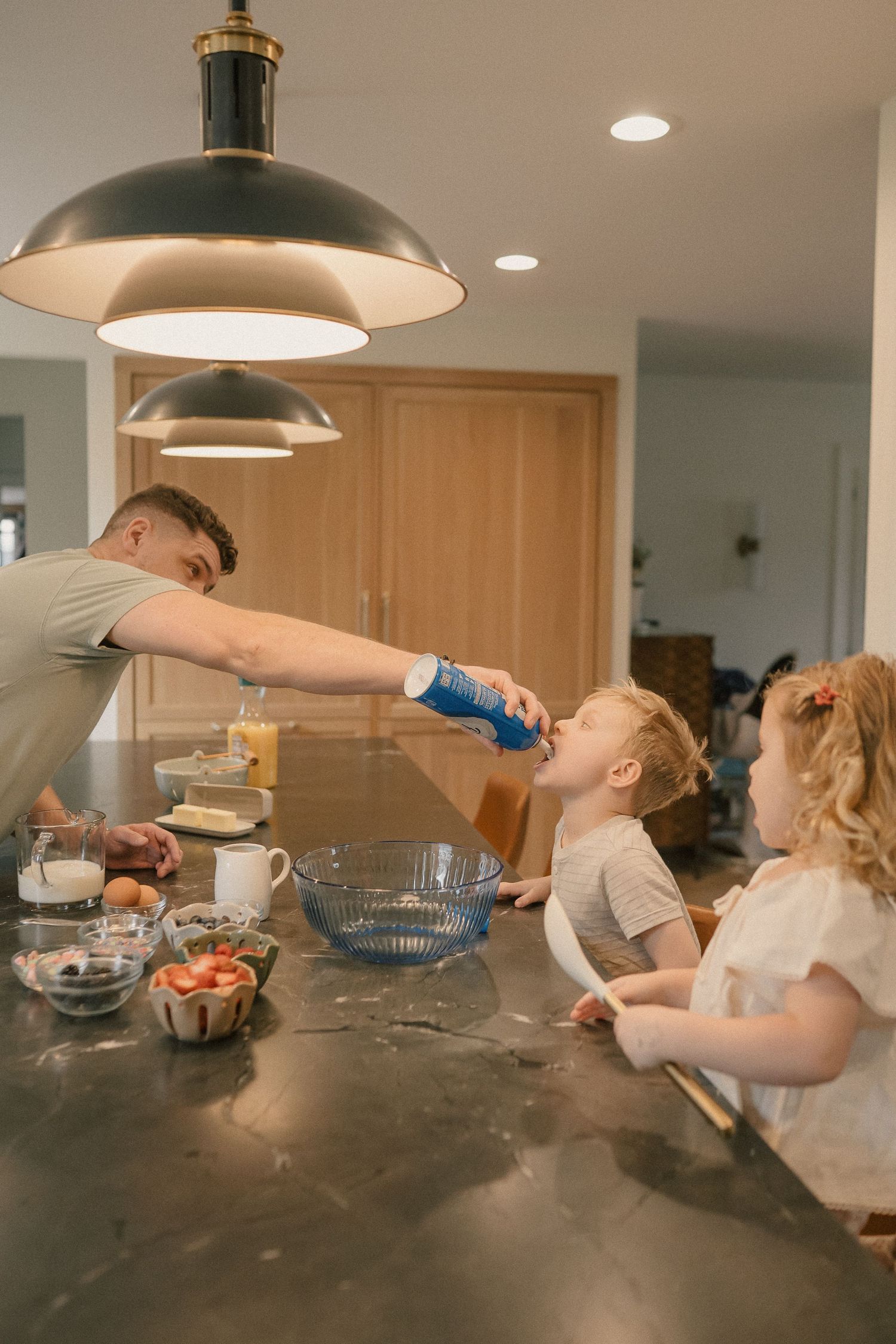 Father giving child a drink while two kids sit at kitchen counter with baking ingredients.