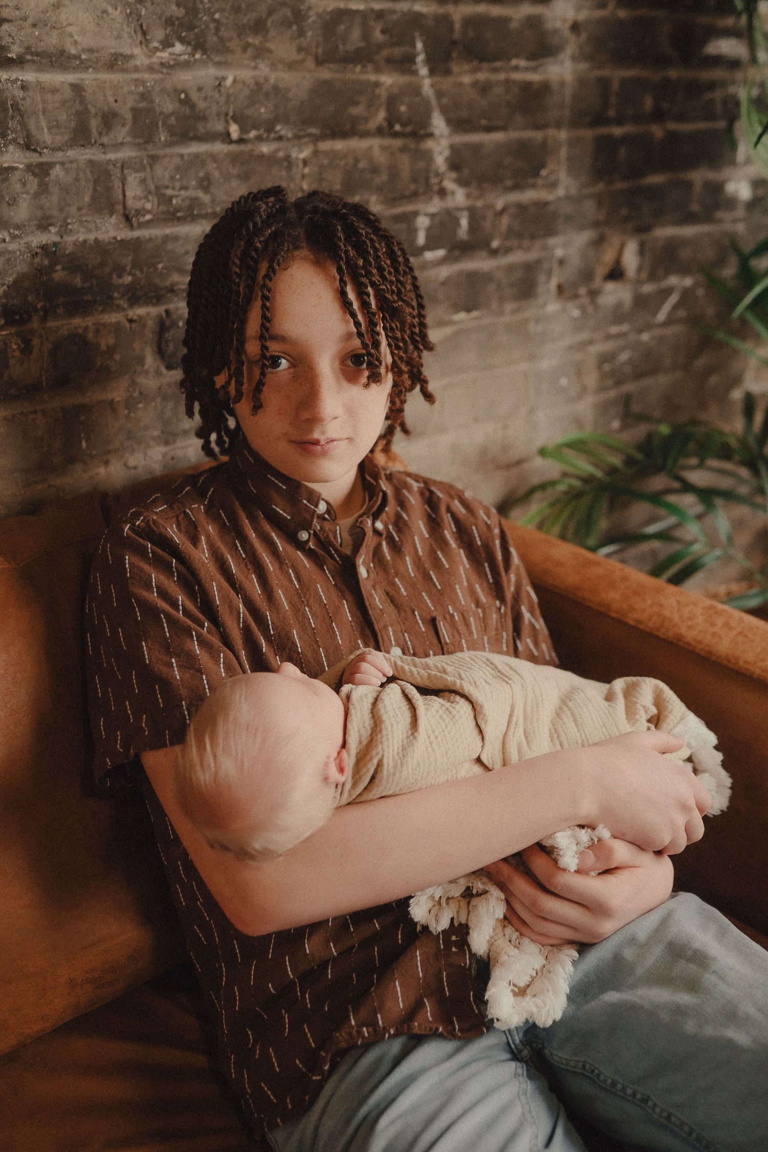 Young person with braided hair holding a sleeping newborn baby while seated in a leather chair near a brick wall.