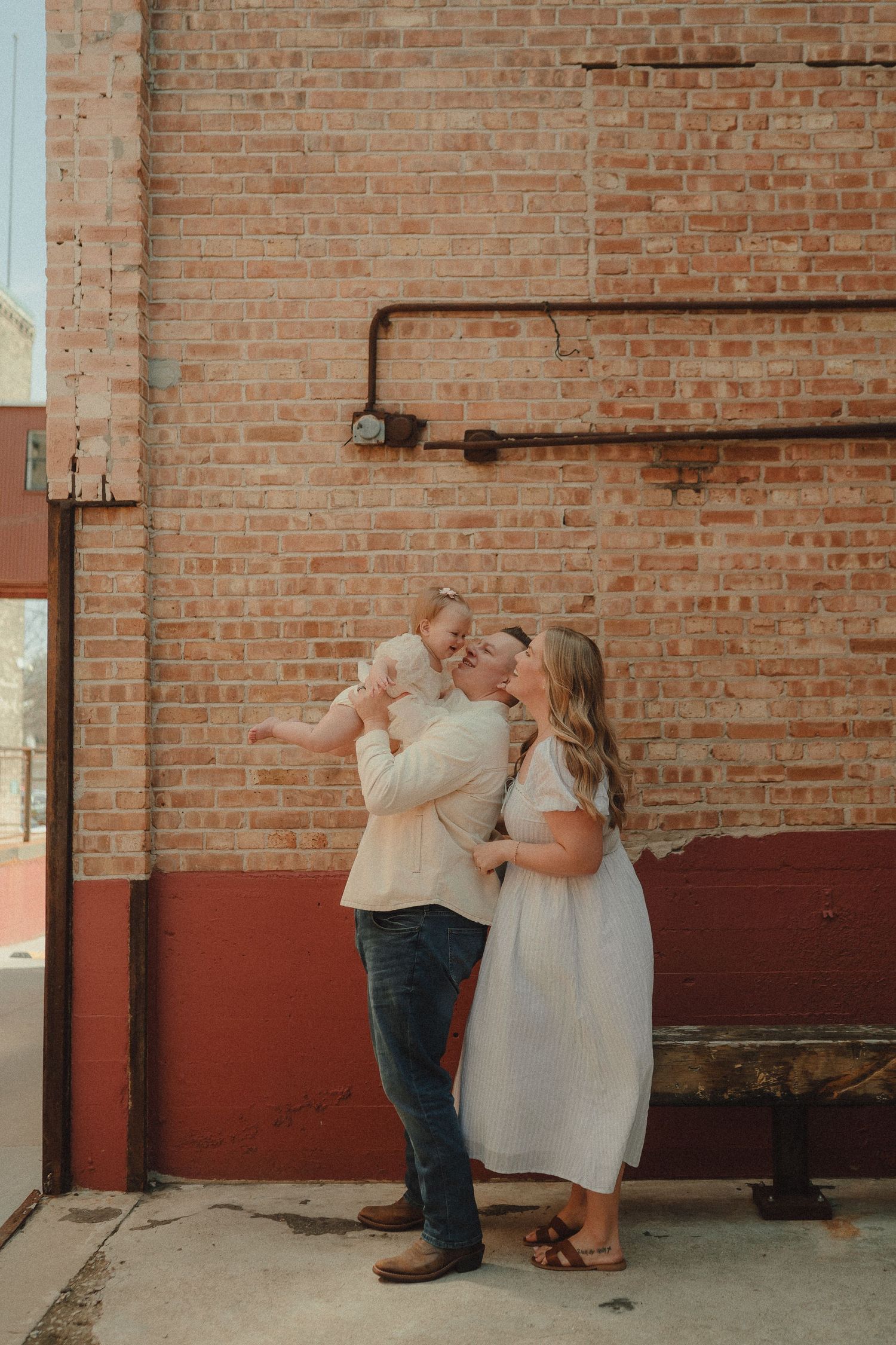 Laughing family lifts smiling baby in the air against a red and brick wall during outdoor portraits.