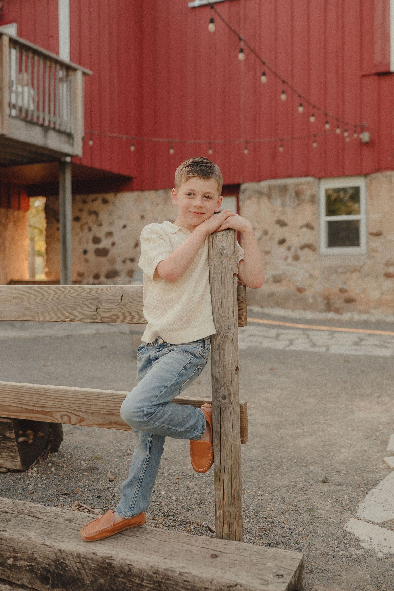 Smiling boy in cream polo and jeans leans on wooden fence post near red barn building.