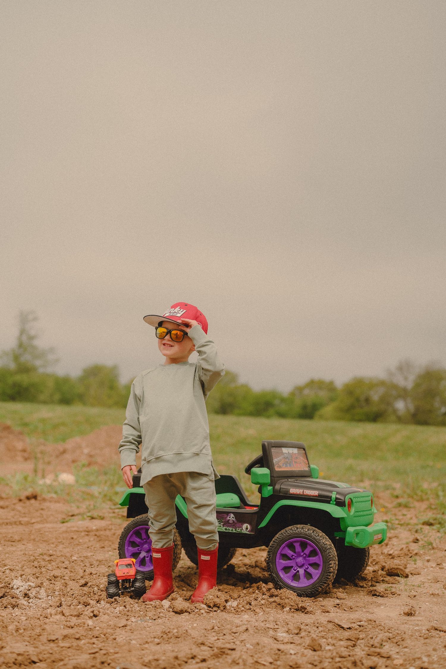 Young boy in red cap and sunglasses stands beside a green toy electric jeep in a muddy field.