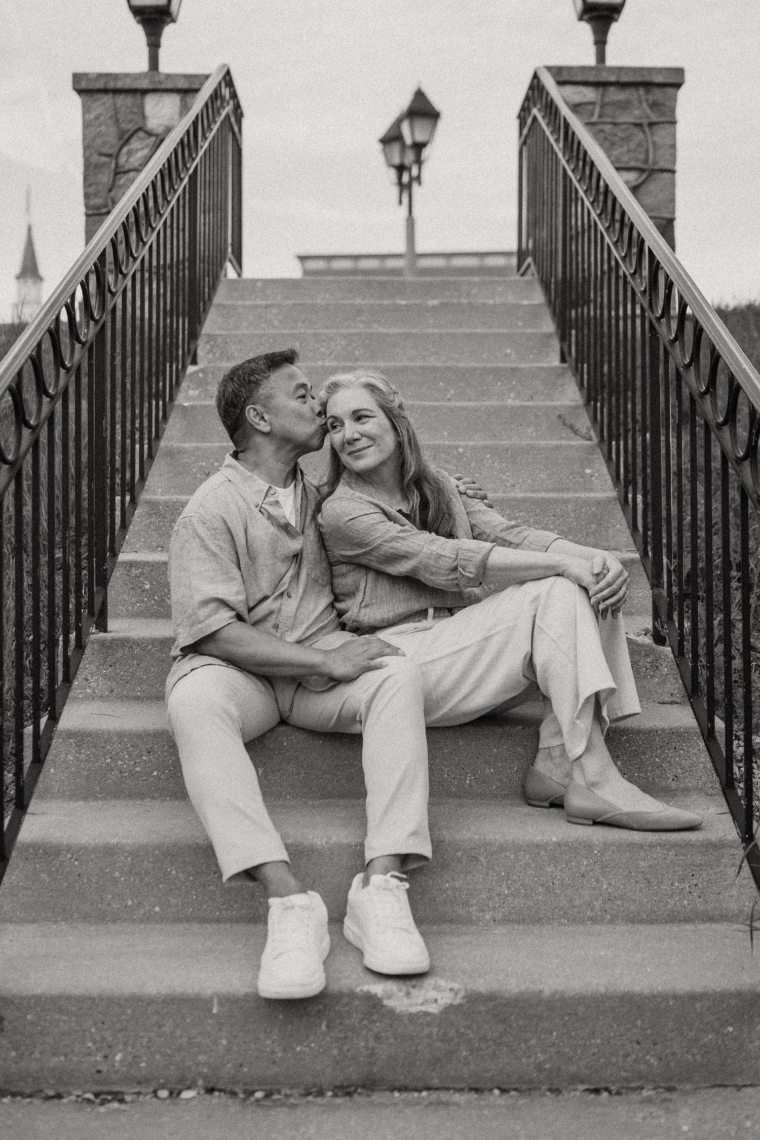 Black and white photo of a couple sitting on steps, sharing a kiss outdoors.