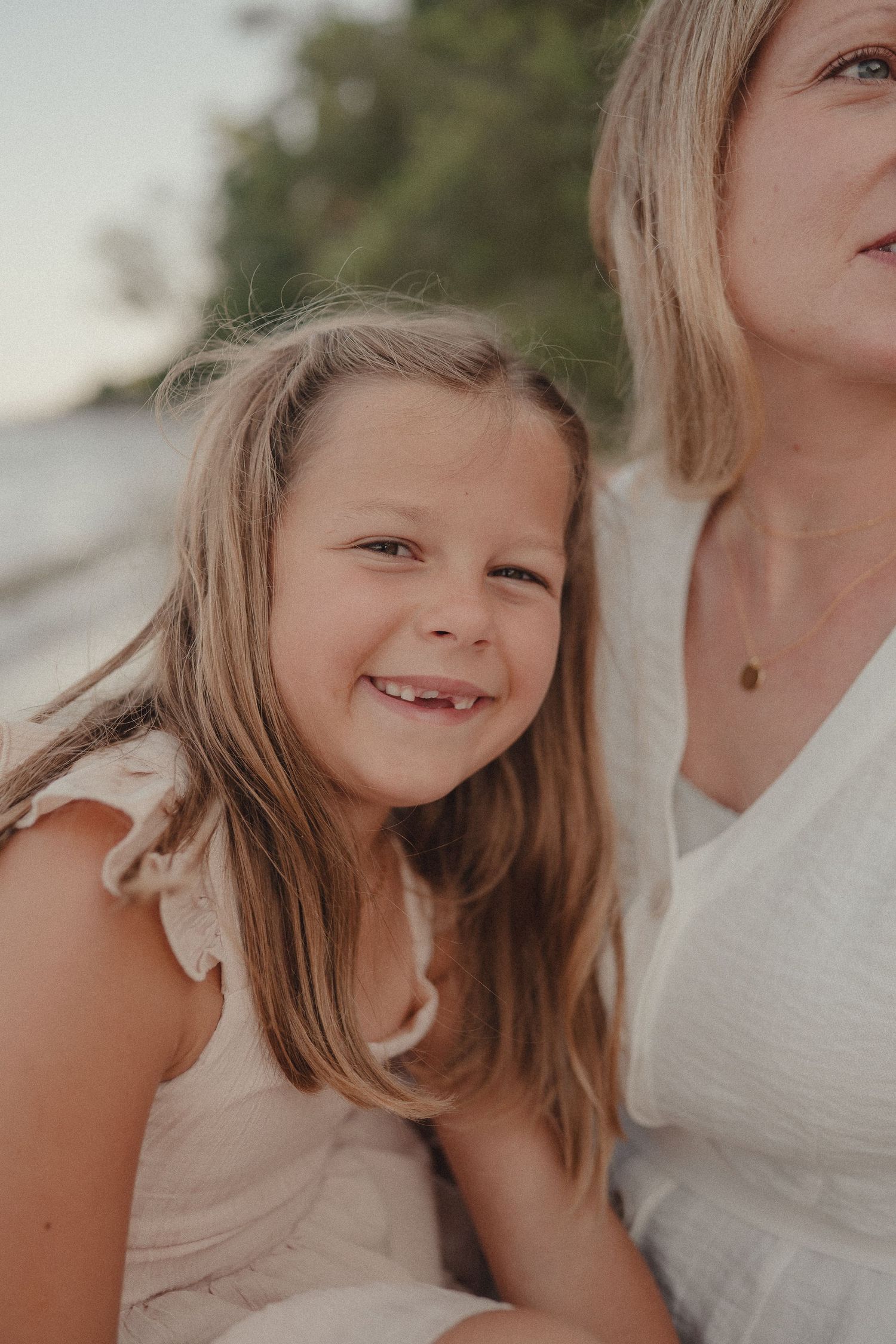 Adorable young girl with long blonde hair smiles brightly outdoors near beach beside a woman in white dress.
