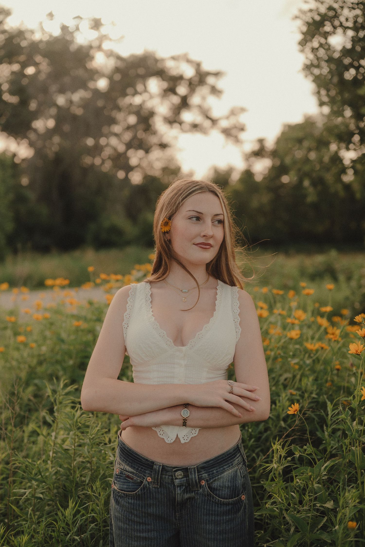 Young woman in white crop top and jeans stands in golden hour flower field with arms crossed.