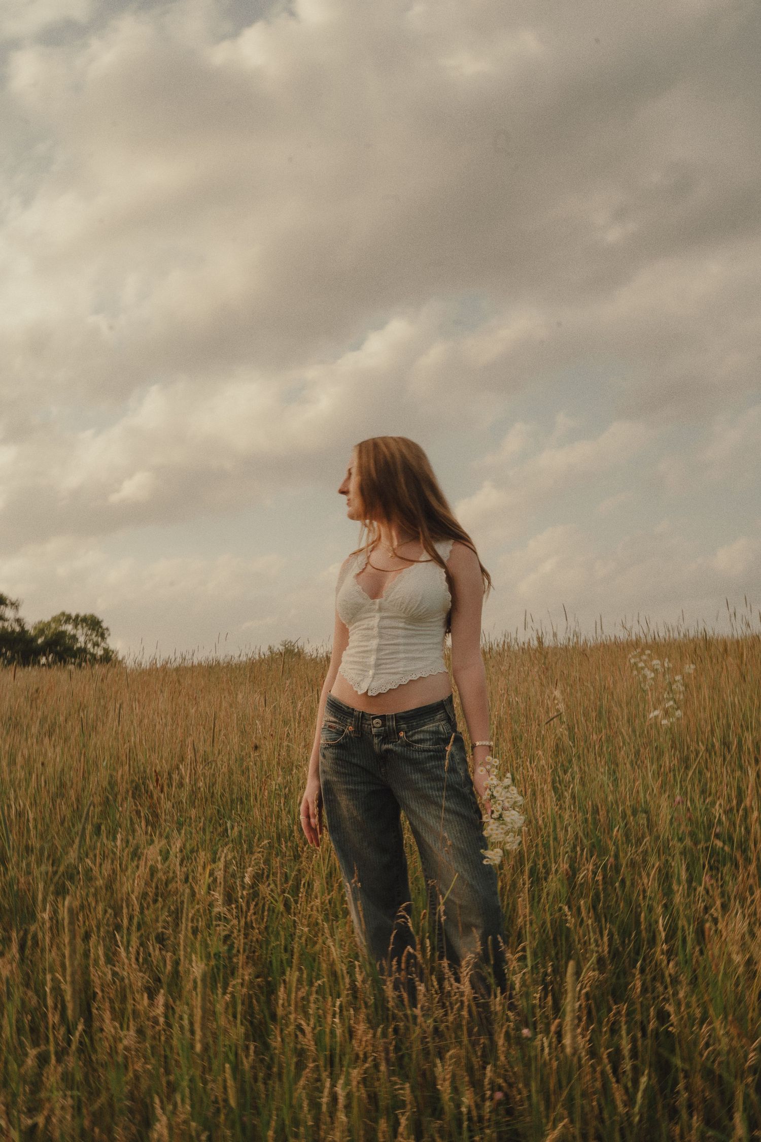 Young woman in white top and jeans standing in a golden field under dramatic cloudy sky.