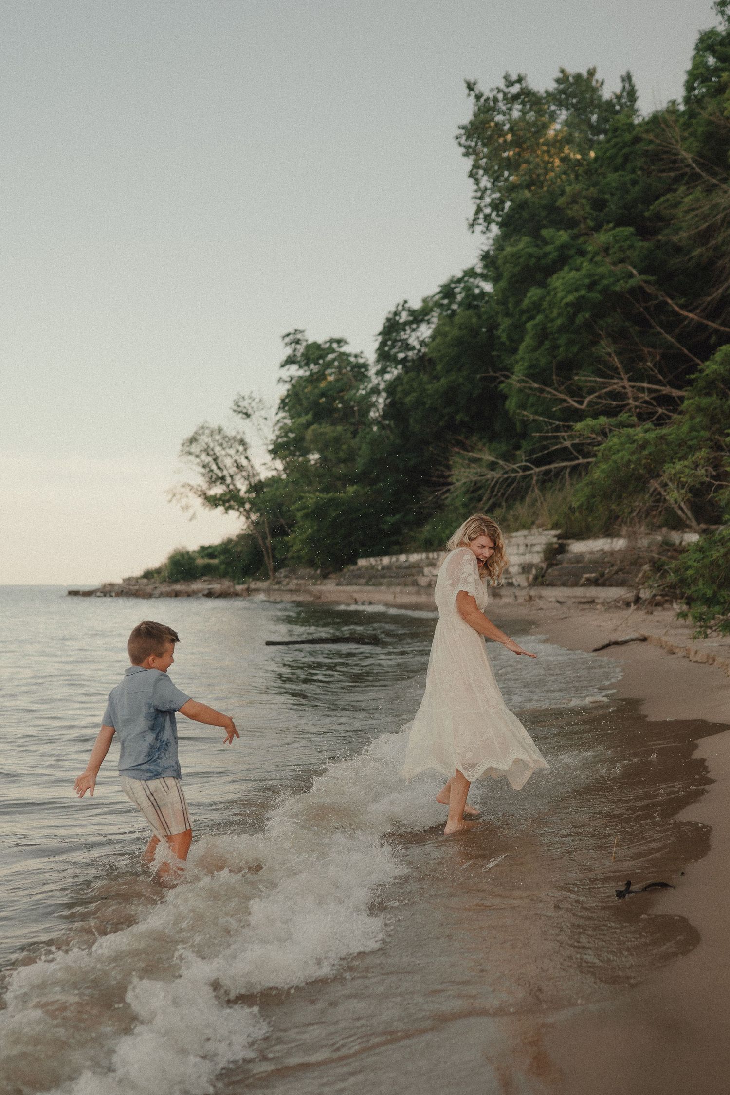 A woman in a white dress and a young boy splash and play along a sandy shoreline near green trees.