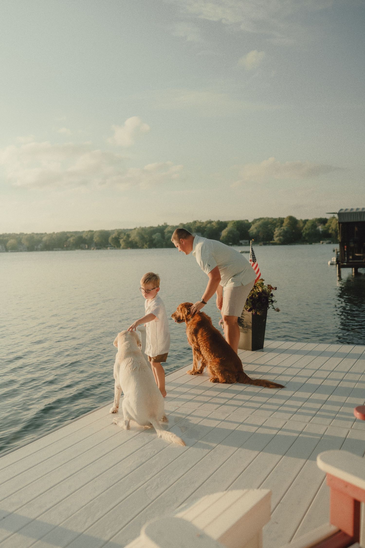 A man and child interact with two dogs on a white dock overlooking a calm lake.