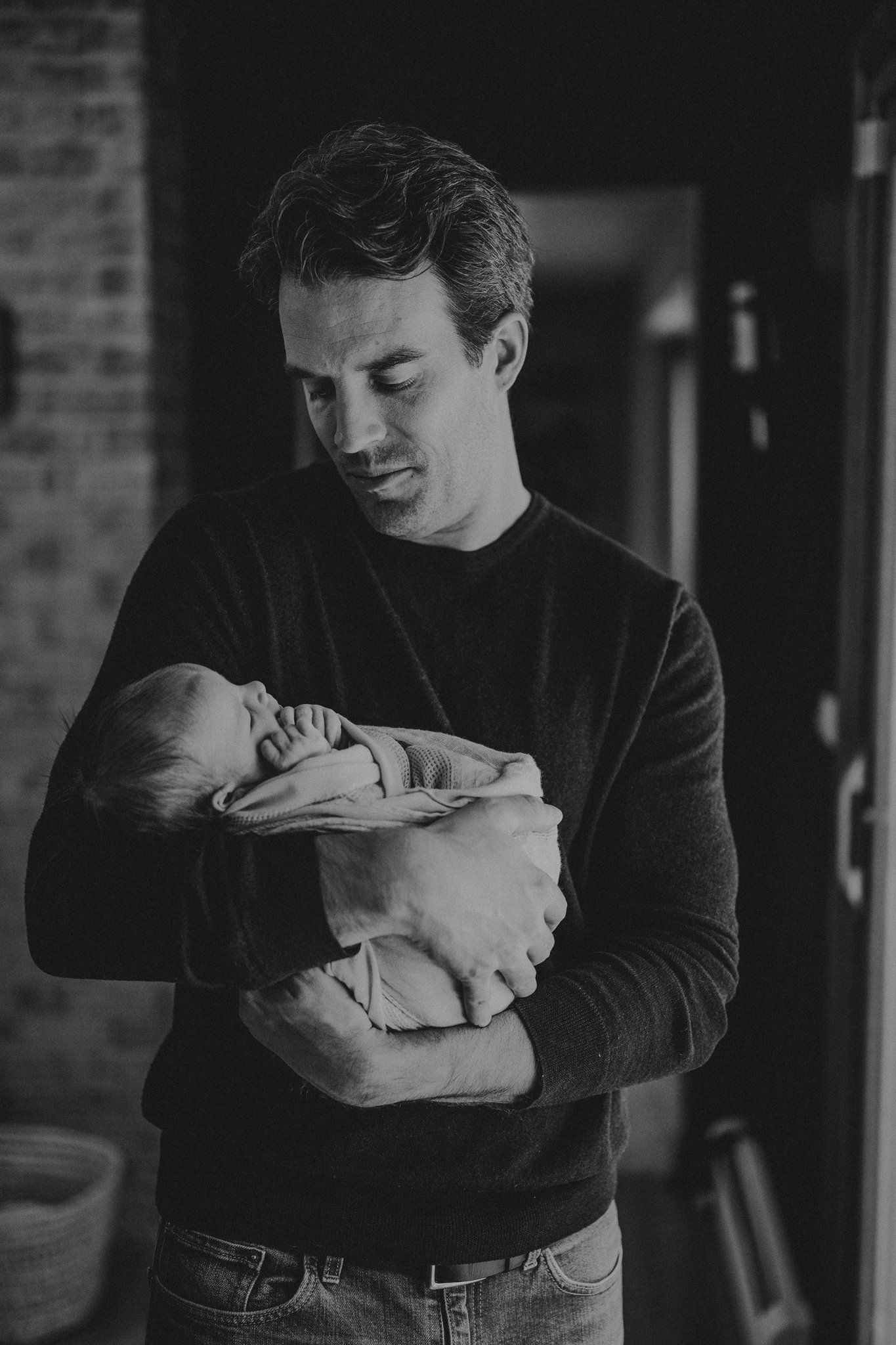 Black and white portrait of newborn being held against exposed brick wall.