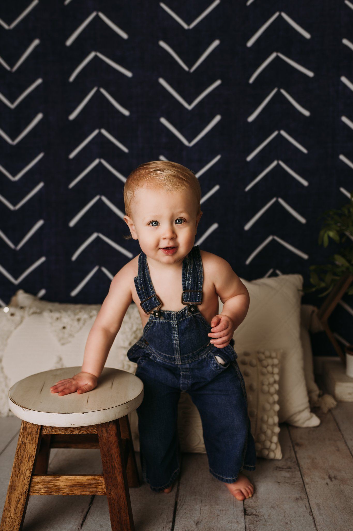 A toddler in denim overalls stands next to a wooden stool against a black and white chevron backdrop.