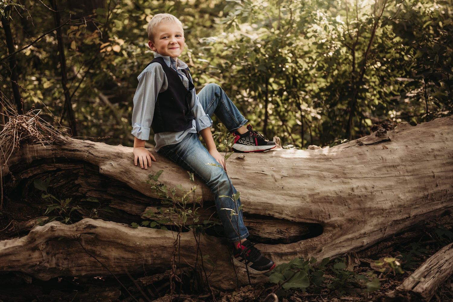 A young person in vest and jeans sits casually on a fallen tree trunk in the woods.