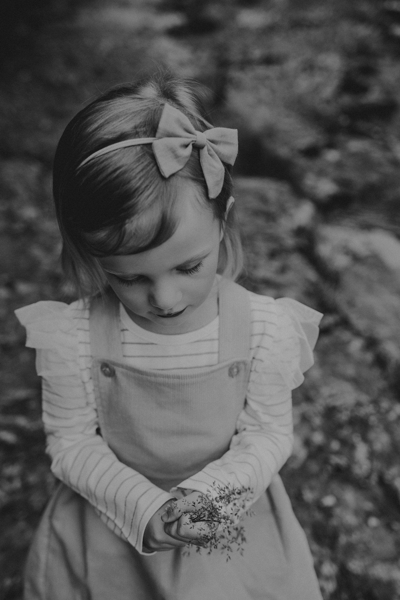 Black and white portrait of a child in overalls and bow looking down at flowers.