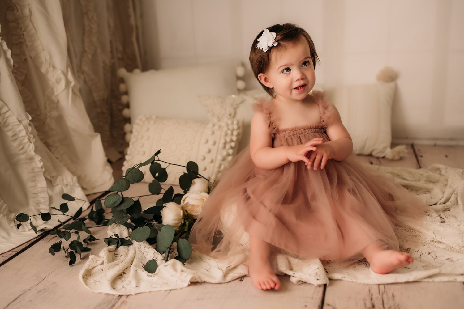 Baby in dusty rose tulle dress on cream textured blanket with white roses and greenery decor.