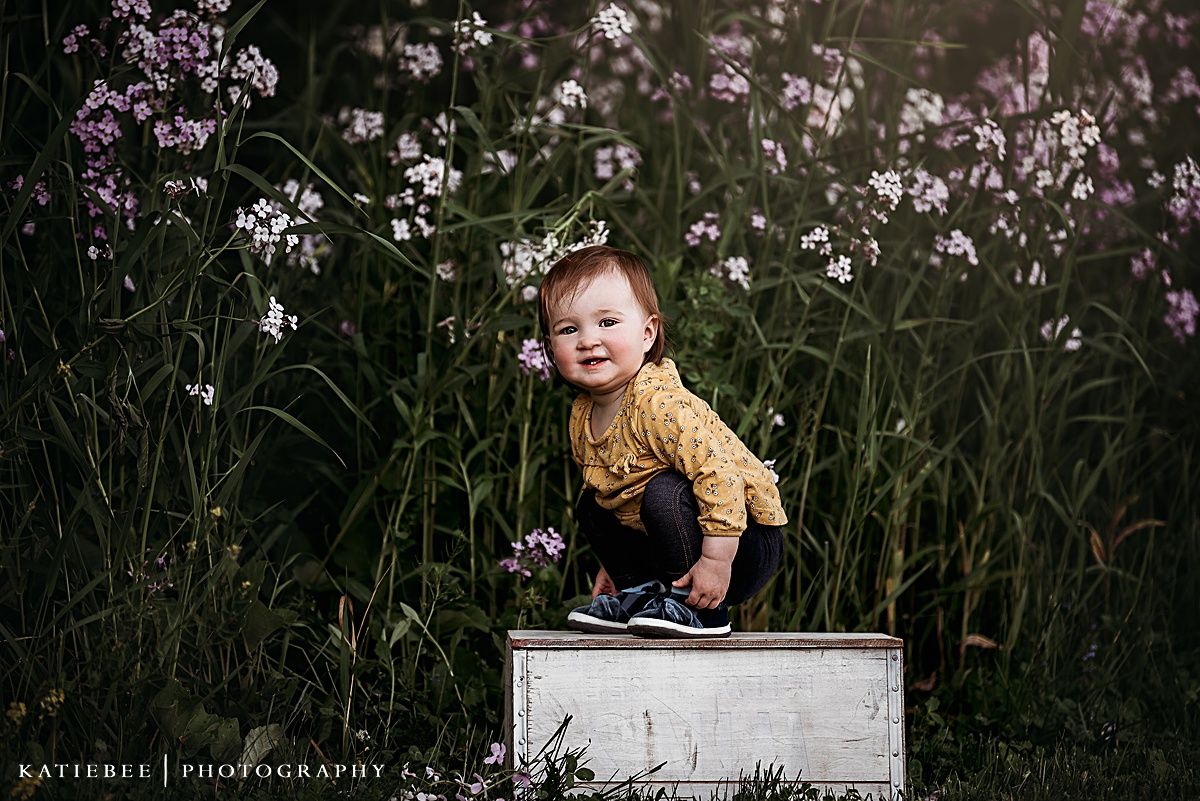 Small child in yellow sweater climbing on white box among tall wildflowers.