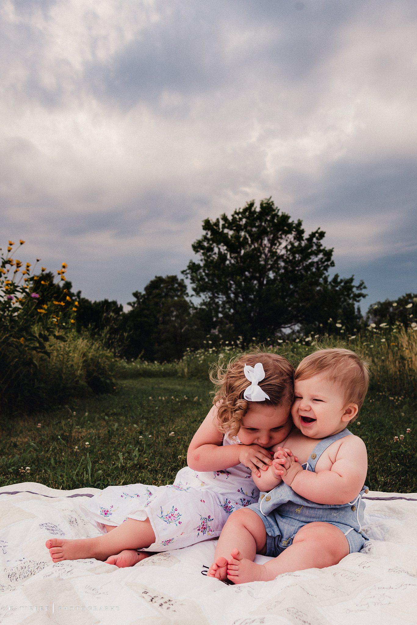 Two babies sit together on a white blanket surrounded by wildflowers and grass.