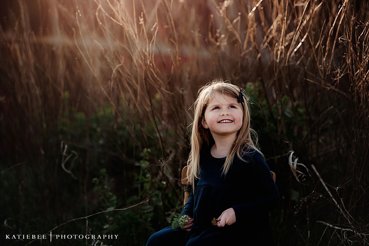 Young child smiling while sitting among tall grasses at sunset.