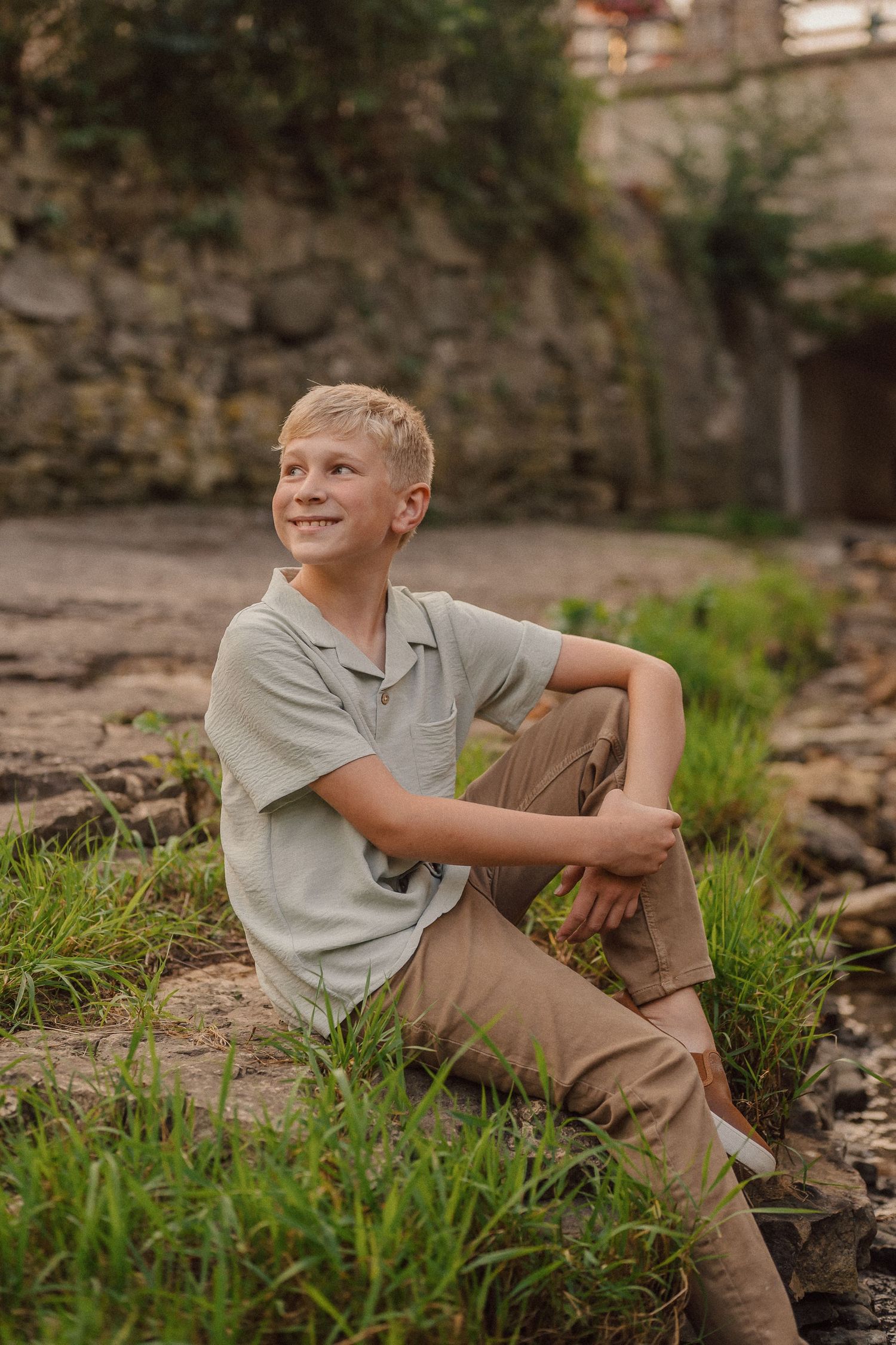 Young child sits on rocky terrain near stone wall wearing gray shirt and khaki pants on sunny afternoon.