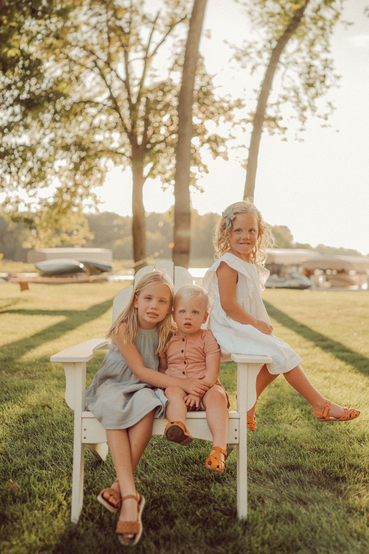 Three young children sit and play on a white bench in a sunny garden setting at sunset.