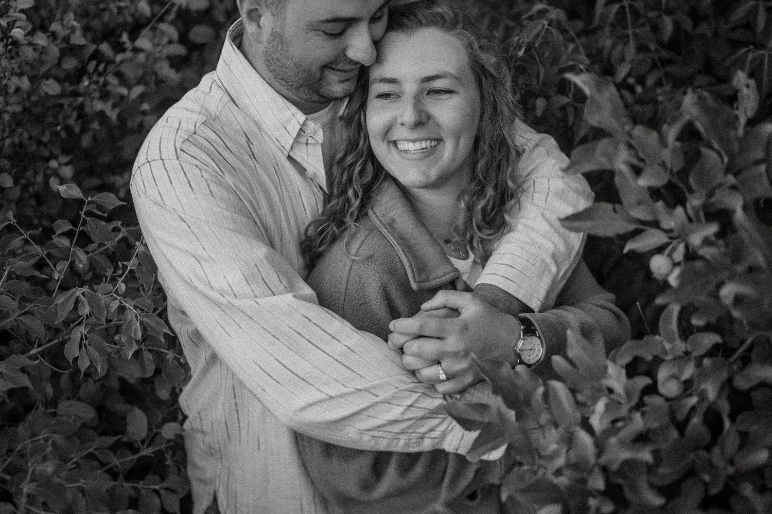 Intimate black and white portrait of couple embracing among leafy branches.
