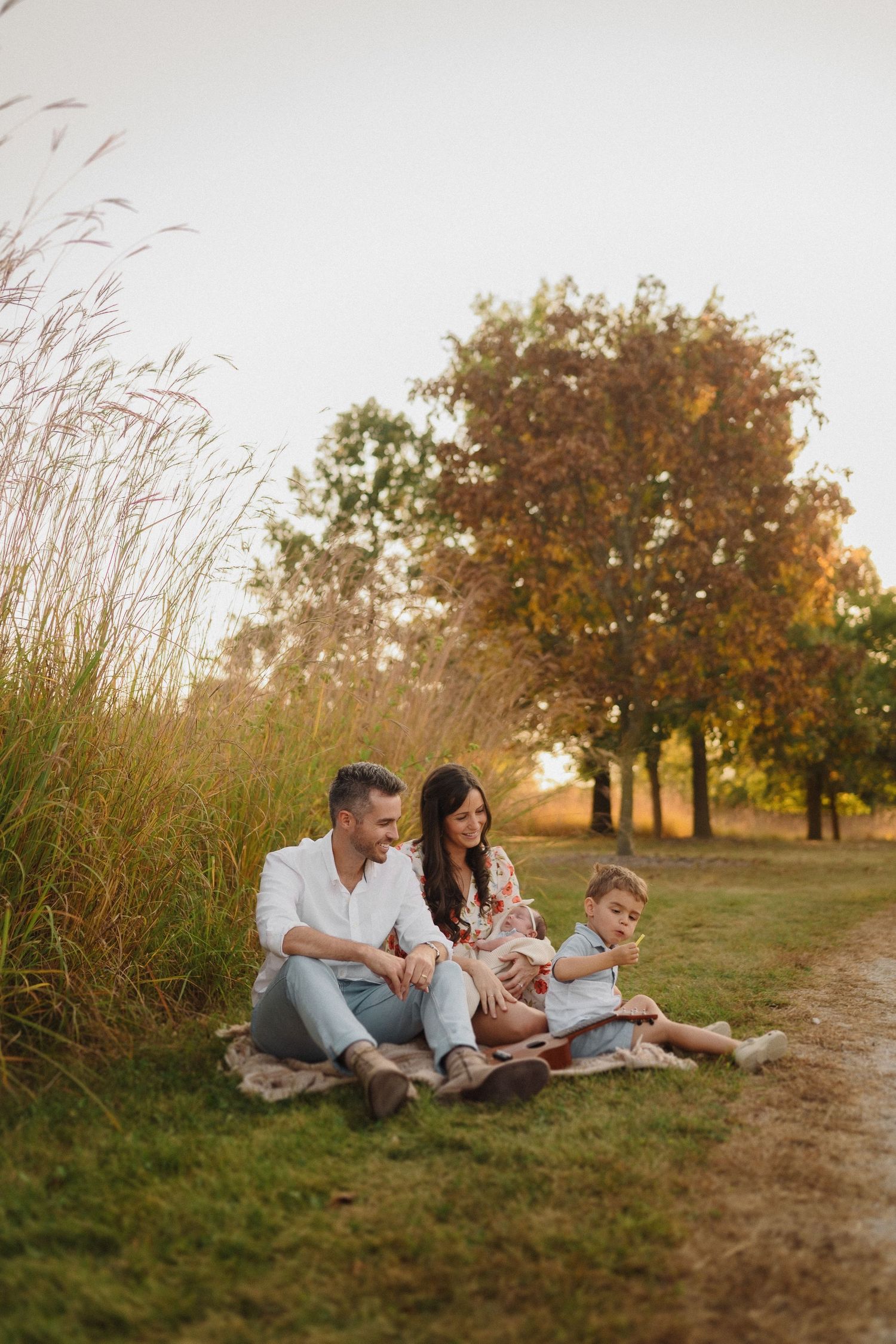 Family sitting together on a blanket at sunset, surrounded by tall grass and autumn trees.