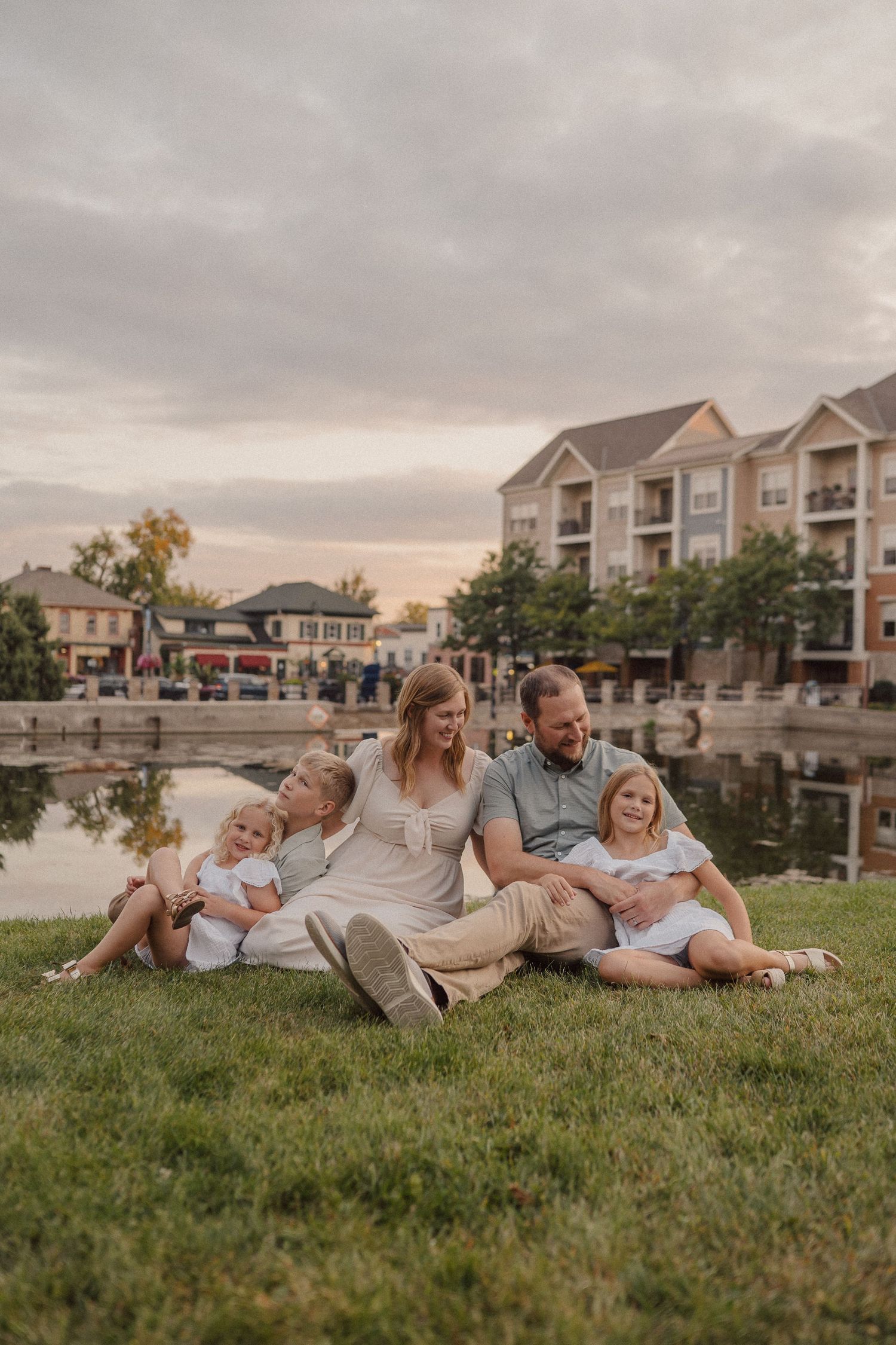 A family relaxes together on grassy lawn near waterfront townhomes during sunset.