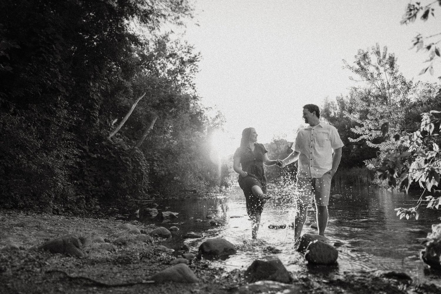 Black and white image of a couple walking among rocks by a river with sunlight filtering through trees.