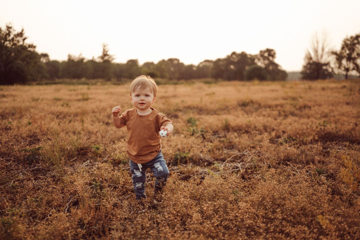 A toddler in brown running joyfully through a golden autumn field.