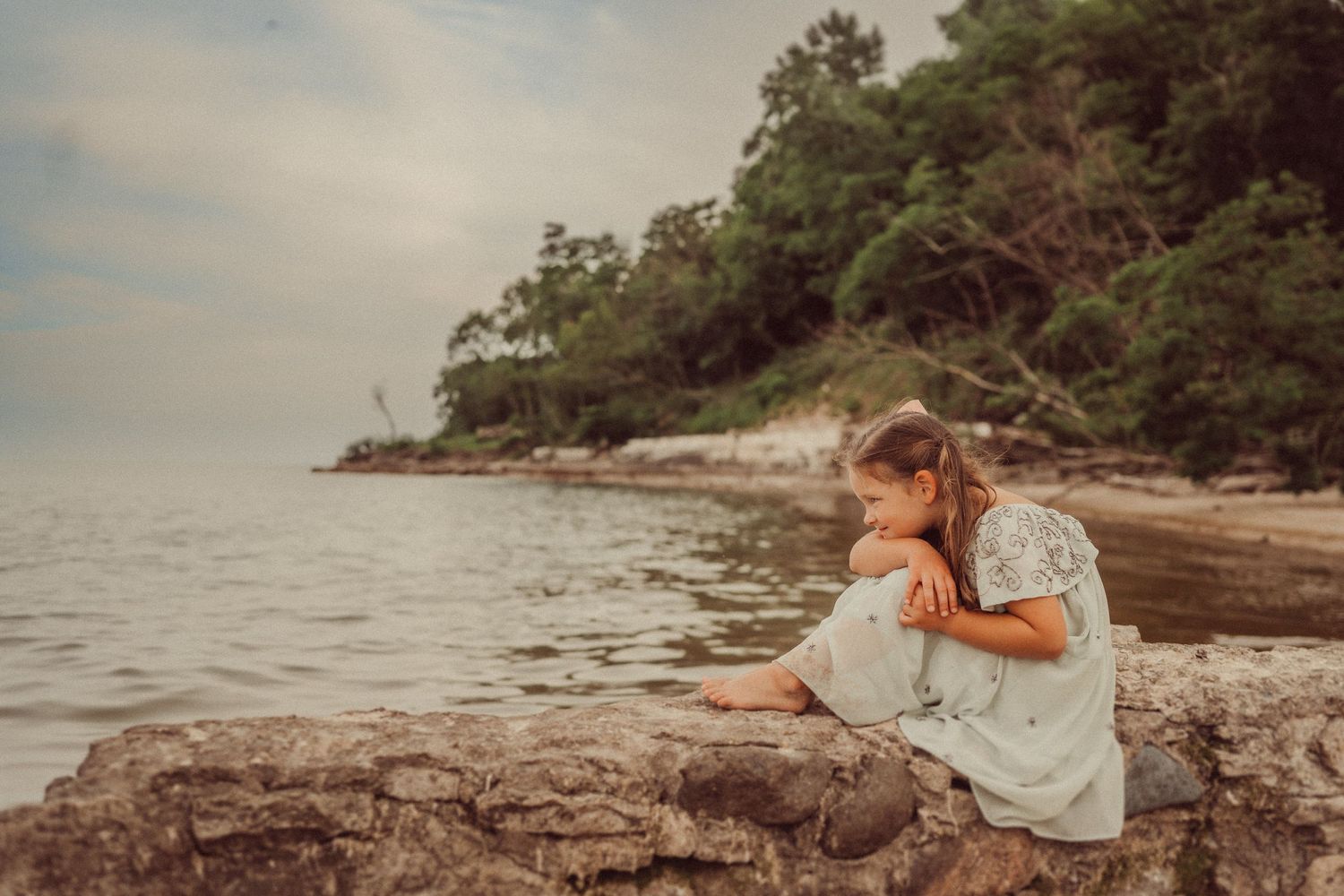 Young explorer in flowing dress sits on rocky beach examining discoveries.