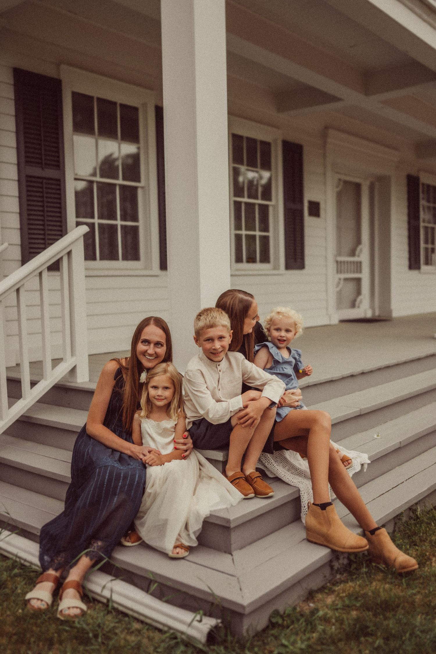 Family gathered on front porch steps of white colonial house with black shutters.