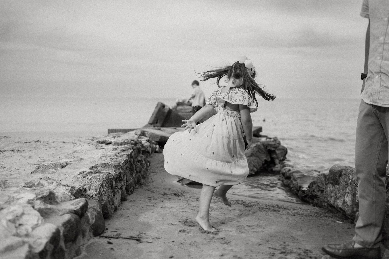A child in a white dress twirls on rocky coastal formations in black and white.