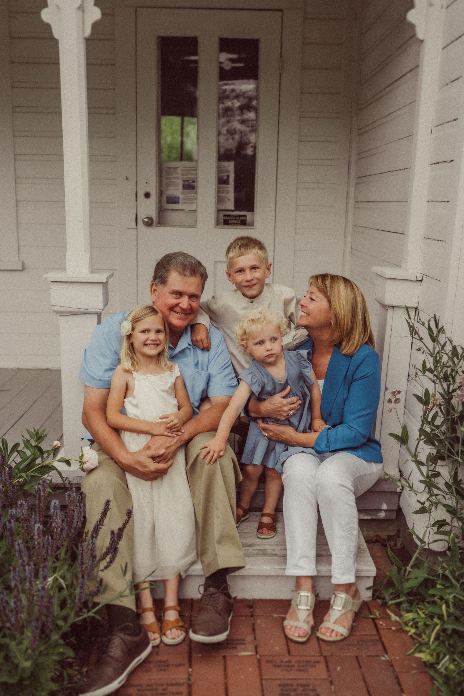 A family sits together on white porch steps surrounded by purple flowers and greenery on a brick walkway.