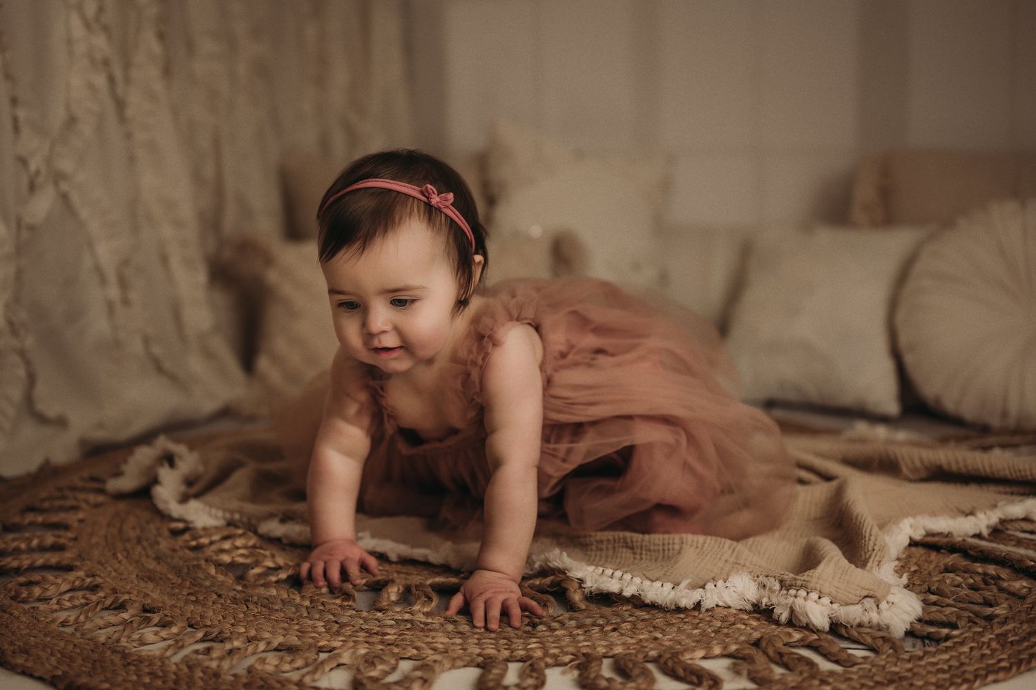Small child in mauve dress crawling on woven rug during indoor photo session.