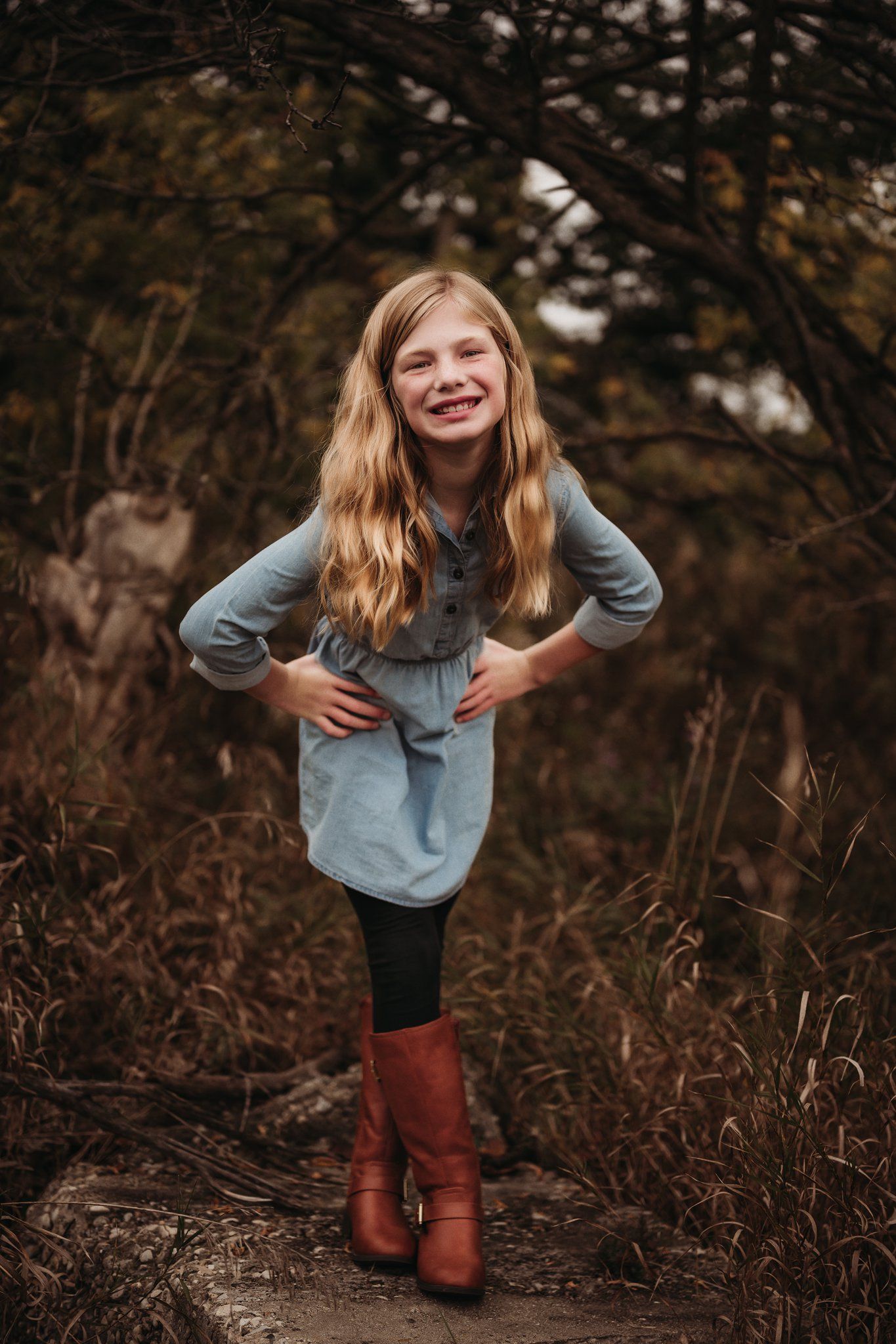 Young person in blue dress and brown boots poses confidently in autumn woods.