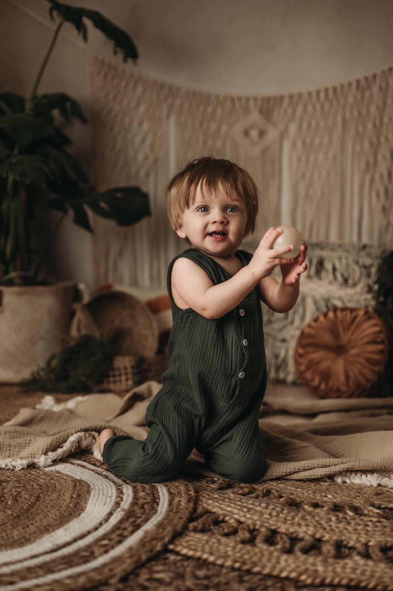 Toddler in green overalls plays with toy on boho-styled rug with plants and macrame decor.