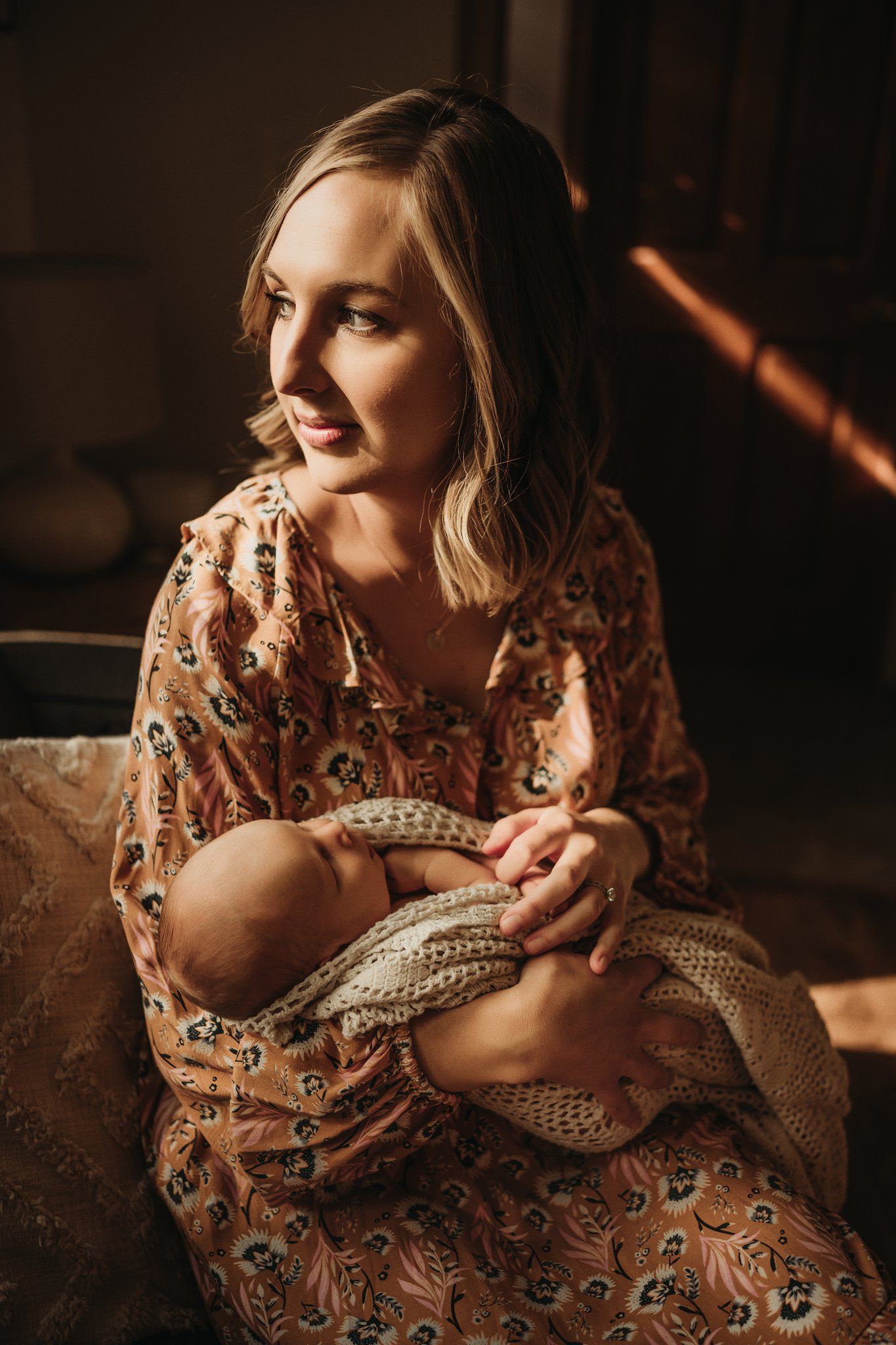 Mother in floral dress gazes lovingly at newborn wrapped in knit blanket.