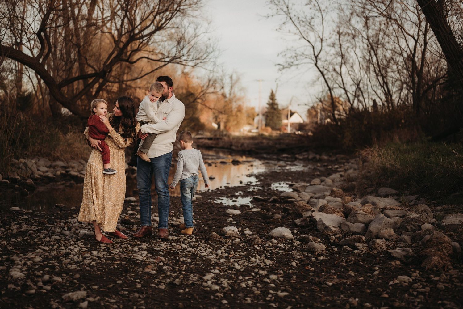 Family photography session on rocky streambed during golden hour with bare trees in background.