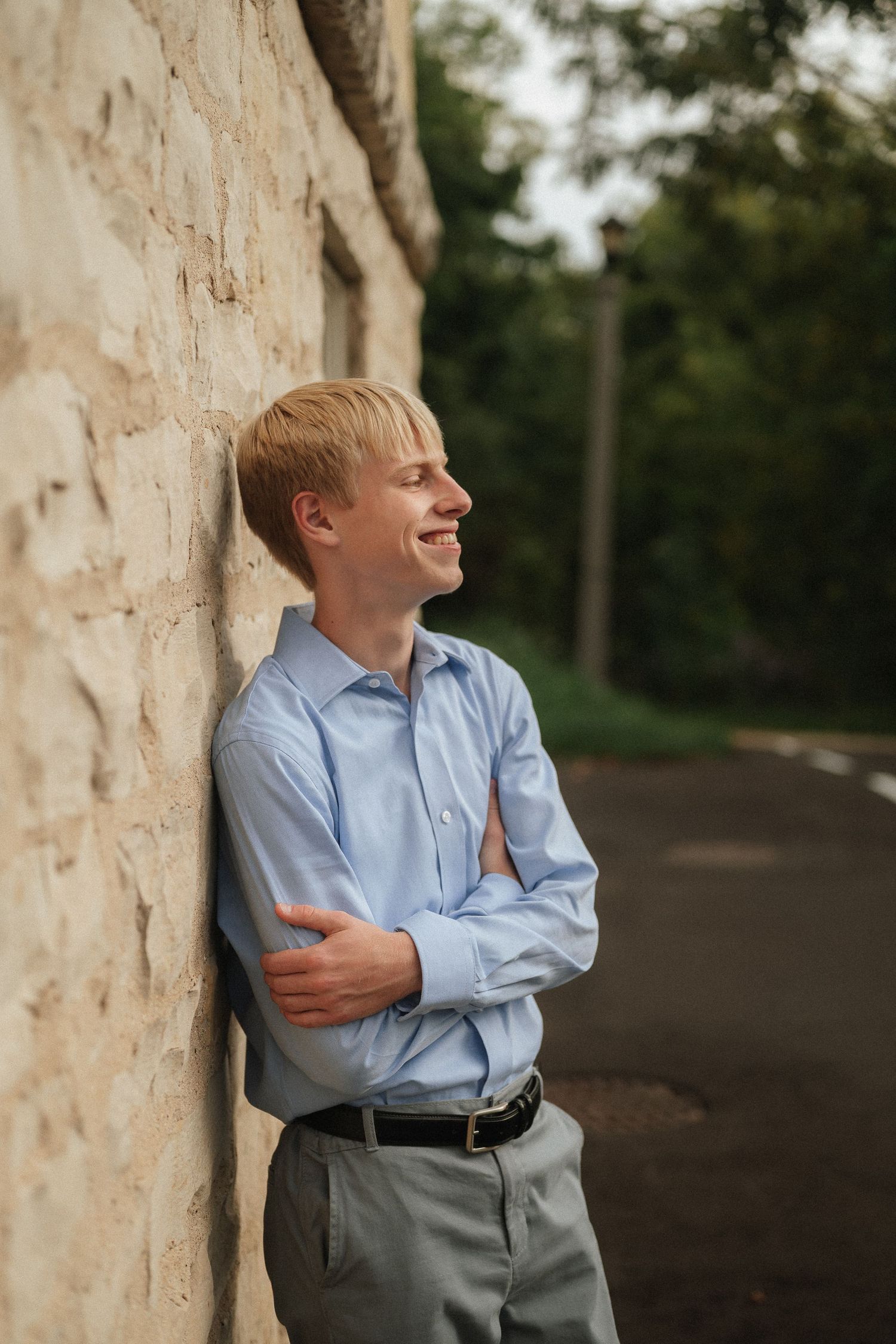 Person in light blue shirt and gray pants leans casually against a textured stone wall outdoors.