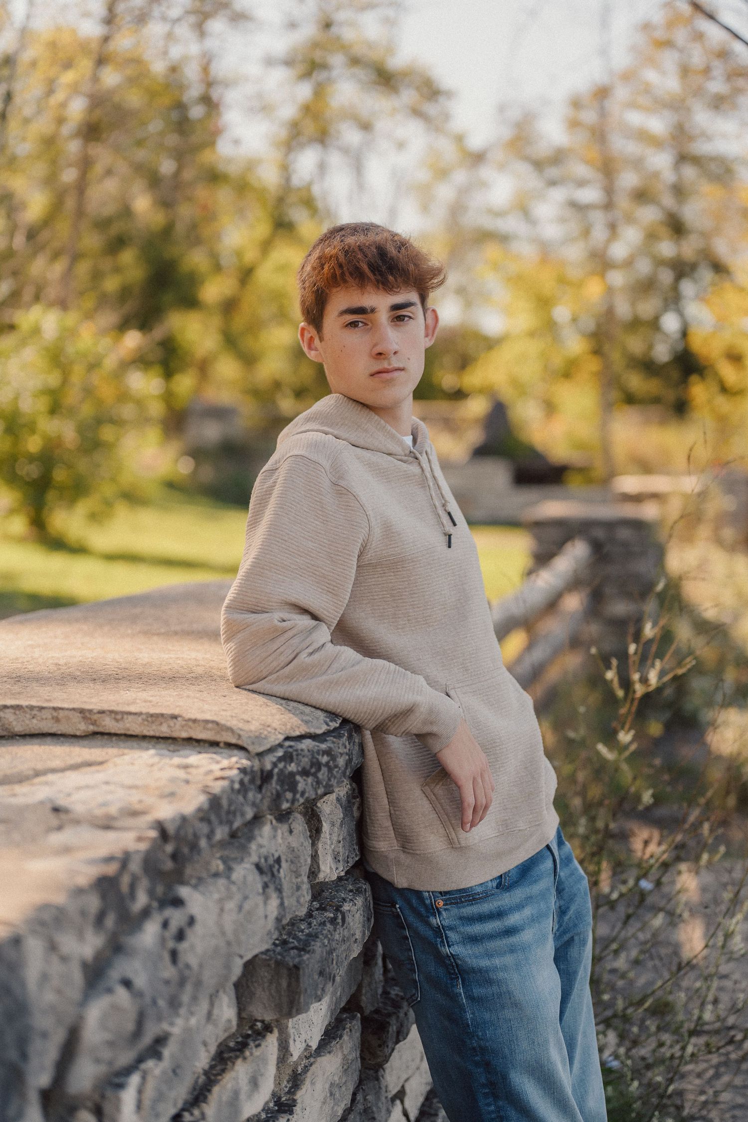 Person in beige sweater leans against weathered stone wall with autumn trees in background.