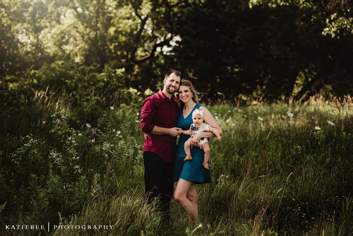 A couple stands together holding their baby in a sunlit meadow with wildflowers and natural greenery.