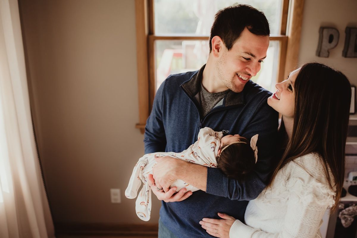 Couple shares intimate moment while holding their newborn by sunny window.