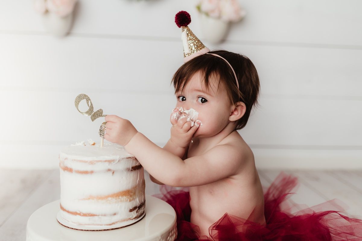 Baby in red tulle skirt reaches for white naked cake during birthday photo session.