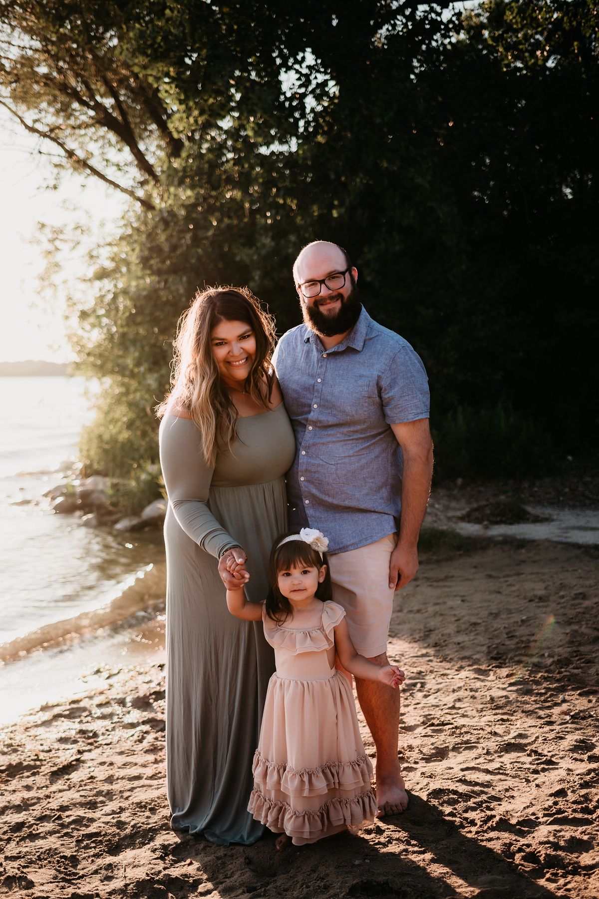 Family of three poses together on a beach at sunset with trees in the background.